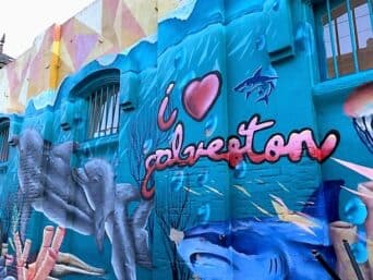 A bright blue mural painted on a brick wall that says "i HEART Galveston"
