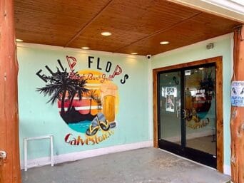 The mural painted on the wall. It says: "Flip Flips Beach Bar & Grill" at the top. The middle is a circle with Palm trees, a frothy beer, and a pair of sandals (flip flops). At the bottom it says Galveston. in Galveston, TX