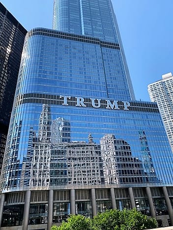 An image of a tall building (skyscraper) with lettering across the front of it: TRUMP. It is a mirrored building, so the reflection of the equally-as-impressive tall building across the street are reflected in it.
