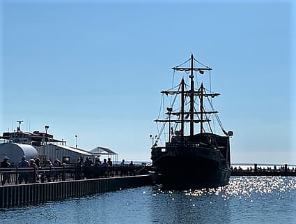 The tall masts of a pirate ship sitting at the dock with no sails. The water in the foreground is blue, while the sky in the background is also blue. The dock and the pirate ship are dark; silhouetted. People are lined up waiting to board the pirate ship ferry to Mackinac Island, Michigan.