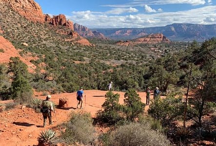 Several hikers moving along a dirt trail. In the distance is the expansive valley surrounded by tall red rocks.