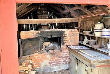 An outdoor building with a hot open oven in the back of it. In the front is a counter which serves as a working space. Shelves on the wall hold bowls and utensils. Amish Villiage.