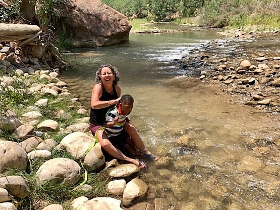 An adult woman with a 6 year old boy sitting on rocks with their feet in the creek. She is trickling water down his neck, and he seems to be objecting!