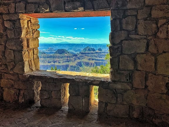 Taken from the inside of a stone building, looking through the window, is the Grand Canyon's North Rim: peaks of grassy mountains inside a low valley (canyon).