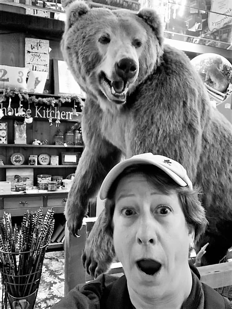 A stuffed and mounted bear is on display inside a store. An adult woman is posing with the bear, so it looks like she's being chased: An expression of shock and surprise is on her face!