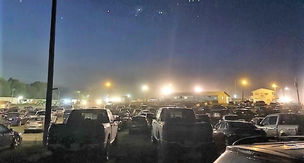 A dark parking lot, full of cars, with floodlights in the background