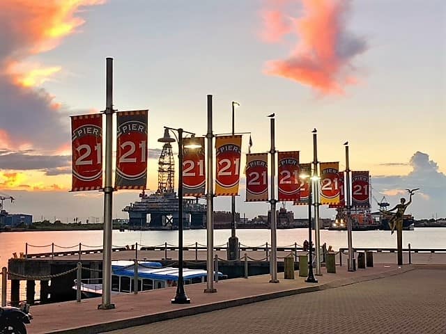 Poles sticking up into the sky, 5 of them spaced apart. Each of them holds 2 banners: one on the left, one on the right., with special hardware. They simply say "Pier 21". In the background, is a sunset happening.