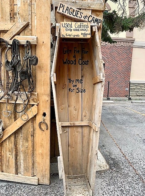 An open coffin, standing on its end, with signs that read, "Used Coffins (used only once)" and "Get Fit For a Wood Coat" and "Try Me on for Size"