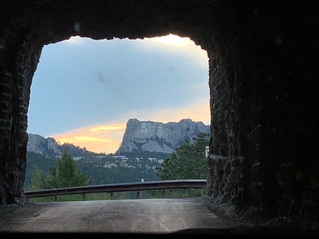 A close up of Mt Rushmore framed by the mouth of a very narrow tunnel. This is one of several tunnels that aligns this way, but you must travel counter-clockwise on the loop! The picture has a dark outer rim, with a bright center. In the middle is Mt Rushmore with pink and yellow highlights in the sky. (Sunset)