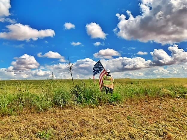 Another shot: It looks like a short stocky man wearing a yellow/orange safety vest and a white hard hat. He's carrying a US Flag that is tattered and torn. He's standing just off the road, heading into/out of an huge open field.