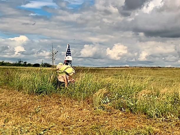It looks like a short stocky man wearing a yellow/orange safety vest and a white hard hat. He's carrying a US Flag that is tattered and torn. He's standing just off the road, heading into/out of an huge open field.