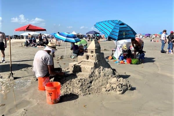 A man, artist Benjamin Guetta, sits at the edge of a pile of sand, sculpting. In the background are several colorful umbrellas and other types of shade awnings, and lots of people milling about.