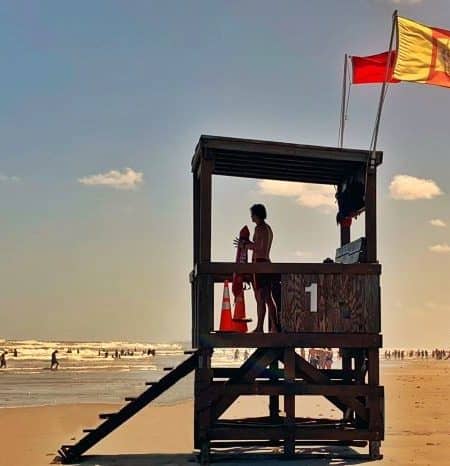 A young man stands attentively at the top of a lifeguard tower, overlooking a crowd of people wading in the Gulf of Mexico on a redflag (no swim) day.