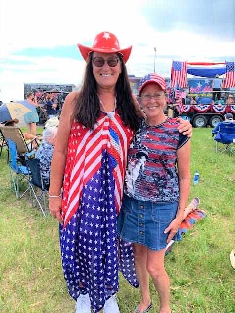 A very tall woman with long dark hair, wearing a red-white-and-blue flag dress, and a red cowboy hat with red stars, stands next to a short woman who is also dressed in red white and blue.