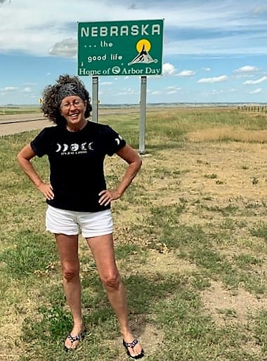 Wearing white shorts and a black tshirt that says, "It's Just A Phase" a woman with big hair stands roadside, arms akimbo, near the Nebraska State Line sign.