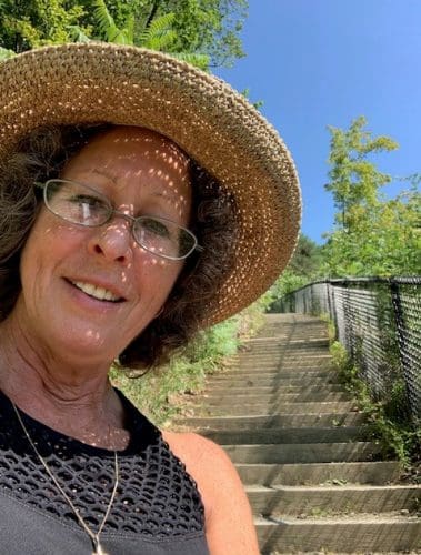 Adult woman wearing a loose-woven straw hat which allows spots of sunlight on her face. There is a steep flight of stairs directly behind her. Home/About Me.